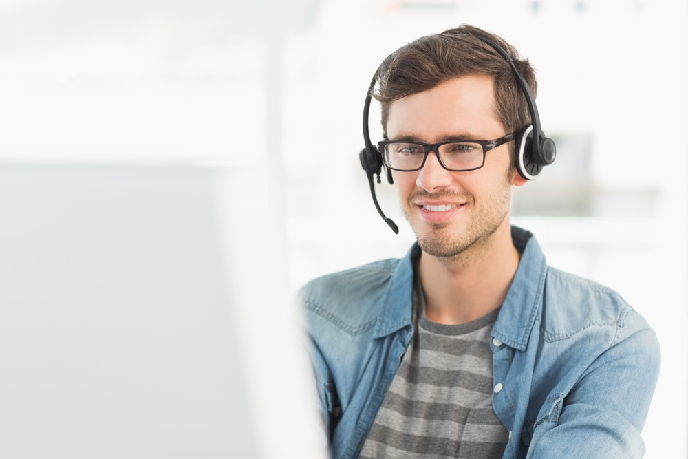 Smiling casual young man with headset using computer in a bright office